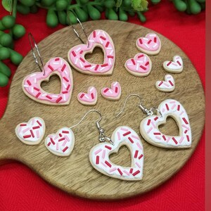 Op de afbeelding: Een set van vier paar hartvormige oorbellen met roze en witte frosting en rode en roze sprinkles. De oorbellen zijn gemaakt van polymer clay en zijn perfect voor Valentijnsdag.