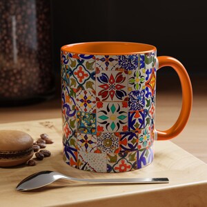 May include: A ceramic coffee mug with a colorful patchwork tile design. The mug has an orange interior and handle. A spoon and a macaron are on a wooden surface next to the mug.