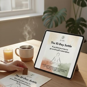 Puede incluir: Una tableta digital muestra "The 10-Day Settle", una guía para el enraizamiento y la restauración. Una taza de café, una vela encendida y un bloque de madera están sobre una mesa de madera. Sale vapor del café.