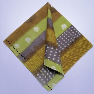 May include: Folded square handkerchief with a geometric design. The handkerchief features stripes of brown, green, and grey, with white polka dots and a scribbled texture. The edges are outlined with black dots and a signature.