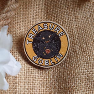 May include: A round wooden pin with a yellow background and black border. The pin features a cartoon goblin holding a red gem and the text "TREASURE GOBLIN".