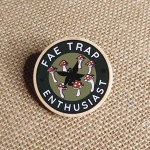 以下が含まれることがあります： 「FAE TRAP ENTHUSIAST」の白い文字が入った丸い木製ピン。中央には黒いシルエットと、赤と白のキノコのイラストが緑色の背景に描かれています。