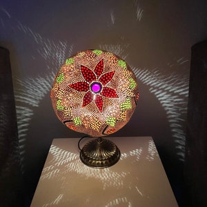 Könnte beinhalten: Eine runde, dekorative Lampe mit einem floralen Design. Der Lampenschirm ist aus einem natürlichen Material gefertigt und zeigt eine rote Blume mit einem rosafarbenen Zentrum. Die Lampe steht auf einem goldenen Metallfuß.