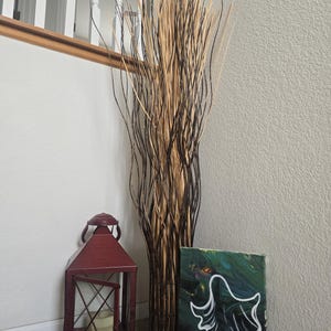 May include: A decorative arrangement featuring tall, dried branches in shades of brown and black. A burgundy metal lantern with an open door and a lit candle sits beside a small painting of a white ghost on a dark green background.