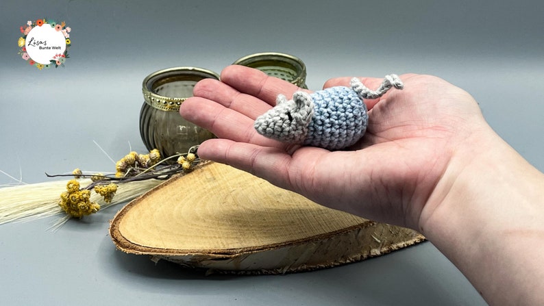May include: A small crocheted mouse toy in gray and light blue with a curled tail. The mouse is sitting in the palm of a hand.