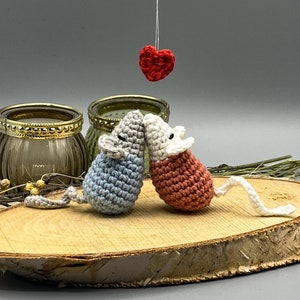 May include: Two crocheted mice in blue and pink are kissing under a red heart hanging from the ceiling. The mice are sitting on a wooden slice.