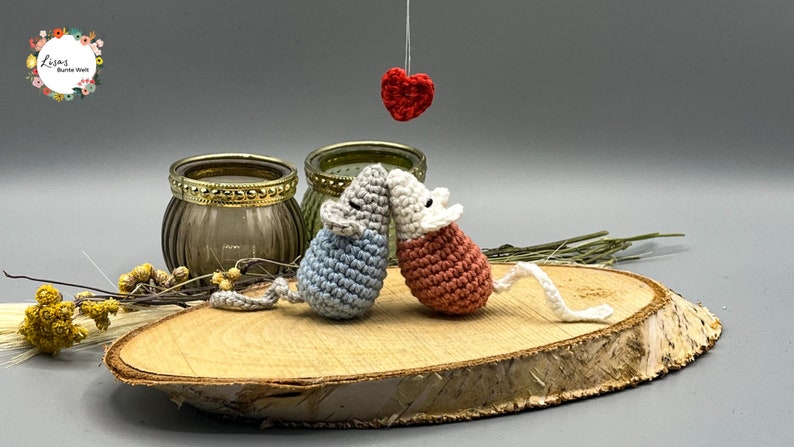 May include: Two crocheted mice in blue and pink are kissing under a red heart hanging from the ceiling. The mice are sitting on a wooden slice.