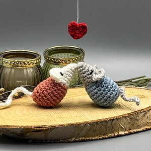 May include: Two crocheted mice in pink and blue are kissing under a red heart hanging from a string. The mice are sitting on a wooden slice with a rustic background.