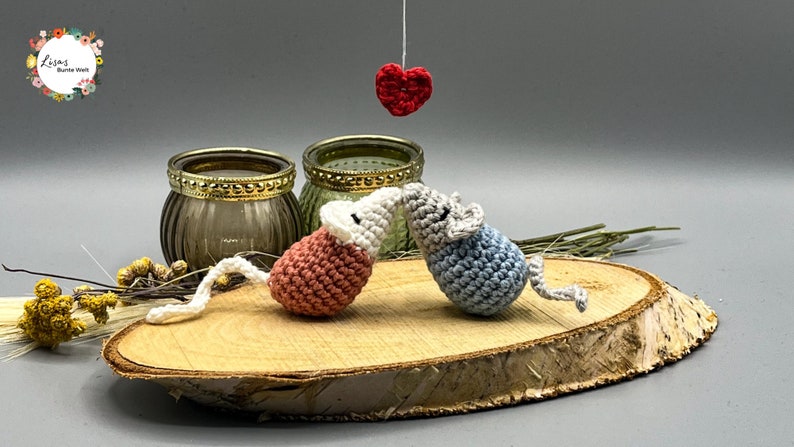 May include: Two crocheted mice in pink and blue are kissing under a red heart hanging from a string. The mice are sitting on a wooden slice with a rustic background.