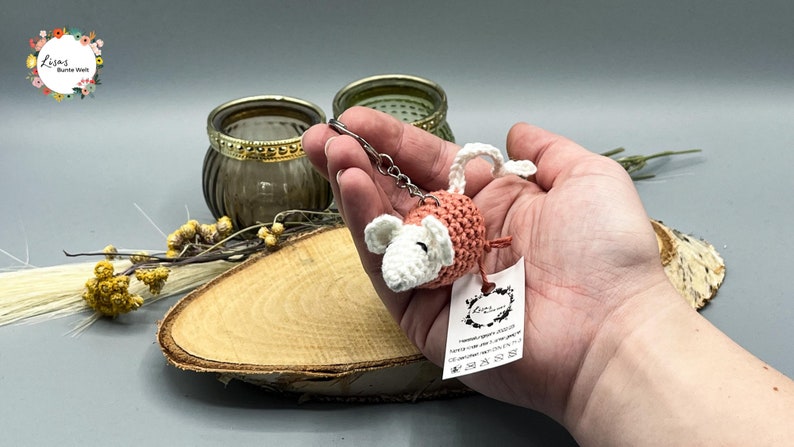 May include: A crocheted pink and white mouse keychain with a silver chain. The keychain is held in a hand with a white tag that says "Lisa's Bunnies World".
