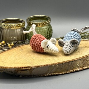 May include: Two crocheted mice, one pink and white, the other blue and gray, are sitting on a wooden slice. The mice are handmade and have small black eyes.