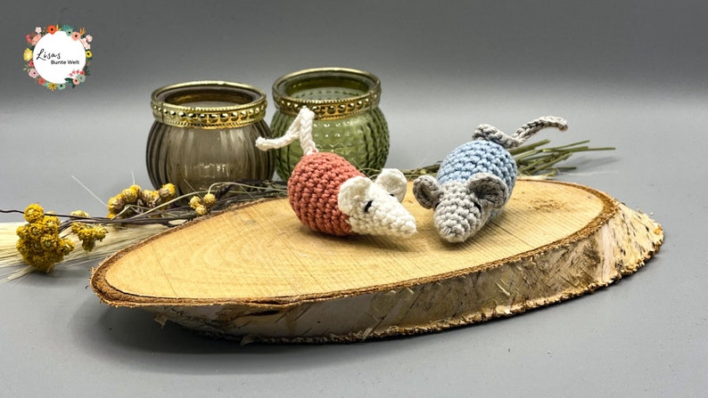 May include: Two crocheted mice, one pink and white, the other blue and gray, are sitting on a wooden slice. The mice are handmade and have small black eyes.