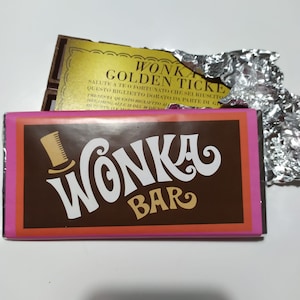 Puede incluir: Una barra de chocolate marrón con un borde rosa y naranja. La barra tiene un logotipo Wonka blanco con un sombrero de copa y el texto "Wonka Bar". El envoltorio tiene una pegatina de lámina dorada con el texto "Wonka Golden Ticket".