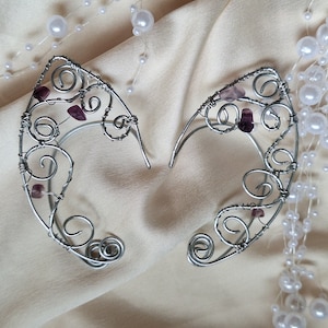 May include: A pair of silver wire ear cuffs with delicate swirls and purple gemstone accents. The cuffs are displayed on a beige fabric background.