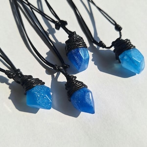 Puede incluir: Cinco collares con colgantes de cristal azul y cordones negros. Cada colgante es un cristal azul facetado, envuelto en un material negro tejido. Los collares se muestran sobre una superficie blanca.