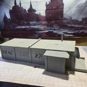 May include: A gray miniature building with a corrugated metal roof, windows, and a chimney. The model has a small attached structure with a ramp. A purple die sits in front of the building.