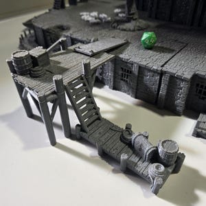 May include: A gray miniature dock with barrels, stairs, and a building. The dock has a wooden plank design and is supported by angled posts. A green die rests on the building's roof. The scene is designed for tabletop gaming.