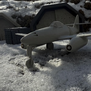 May include: A gray model airplane with a long, narrow fuselage and swept wings. The plane is sitting on a snowy surface, with a concrete structure in the background.
