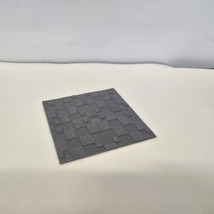 May include: A square, dark gray tile featuring a textured surface with a pattern of raised, unevenly sized squares and rectangles. The design includes subtle, etched details on some of the individual blocks, creating a grid-like appearance.