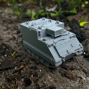 May include: A gray plastic model of a military armored personnel carrier with a turret on top. The model is sitting on a bed of dirt and leaves.