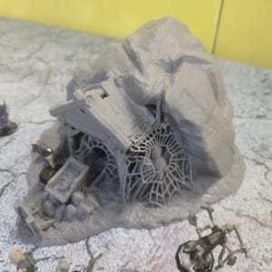 May include: A gray miniature fantasy terrain piece depicting a weathered stone structure with a spiderweb, a small coffin, and a rocky exterior. The miniature is designed for tabletop gaming.