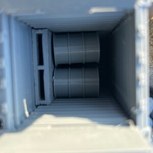 May include: Two gray barrels are stacked inside a gray container with a wooden pallet. The container is open and the barrels are visible.