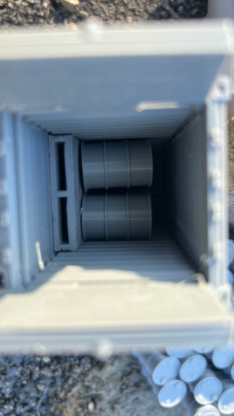May include: Two gray barrels are stacked inside a gray container with a wooden pallet. The container is open and the barrels are visible.