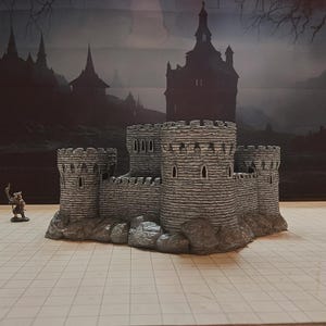 May include: A gray miniature castle with four turrets and stone-like texture. The castle sits on a rocky base, with a small figurine of a warrior standing nearby. The background features a dark, ominous landscape.