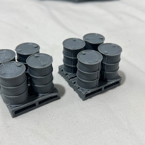May include: Two sets of miniature gray barrels, each set arranged on a small pallet. The barrels are cylindrical with a ridged texture and a lid on top. The pallets have a slatted design. The items are likely for a model or diorama.