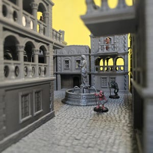 May include: A miniature gray town square scene with buildings, a fountain, and cobblestone streets. Small figurines of fantasy characters are placed throughout the scene, creating a diorama. The background is a solid yellow.