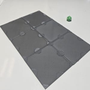 May include: A set of six gray, square game tiles with textured surfaces and corner details. A green, multi-sided die rests nearby. The tiles are arranged in a grid, suggesting a tabletop game setting.