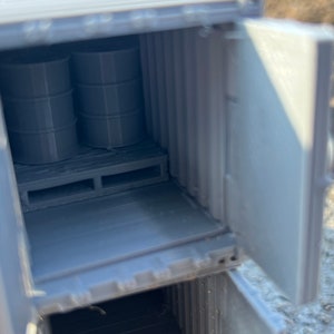 May include: A gray model of a shipping container with an open door. Inside the container are two gray barrels on a wooden pallet.