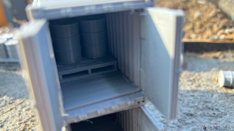 May include: A gray model of a shipping container with an open door. Inside the container are two gray barrels on a wooden pallet.