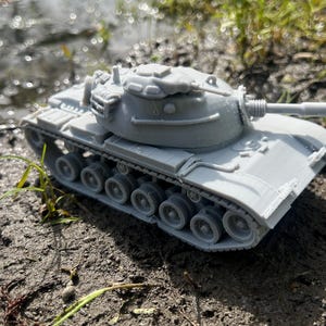 May include: A gray toy tank with a long barrel and detailed tracks. The miniature military vehicle is positioned on muddy ground near water and grass. The tank's turret and body are visible.