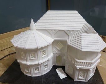 May include: White 3D printed model of a two-story building with a round tower. The building has a steeply pitched roof with a decorative dormer window. The tower has a conical roof and several windows. The model is on a black surface.