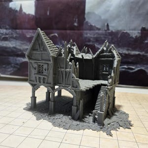 May include: A gray, 3D printed model of a ruined building with a wooden frame and a stone foundation. The building has a collapsed roof and broken windows. The model is set on a gray, textured surface.