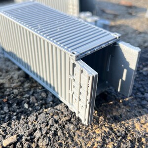May include: A gray metal shipping container with the doors open. The container is sitting on a gray gravel surface.