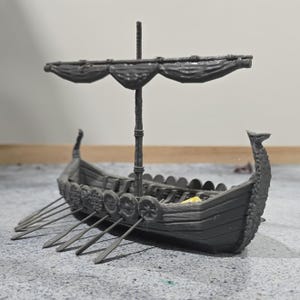 May include: A gray model Viking ship with a mast, sail, and oars. The ship features a detailed hull with decorative shields and a carved prow. The sail is draped, and the oars extend from the sides, suggesting movement.
