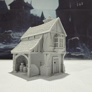 May include: A gray 3D printed miniature model of a small, two-story building with a gabled roof, a stone facade, and a wooden door. The building has a small window on the side and a larger window on the front. The building is set against a blurry background of other buildings.