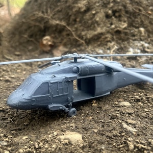 May include: A gray toy helicopter model with a black cockpit and a detailed design. The helicopter is sitting on a dirt surface.