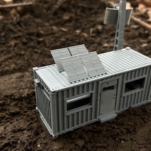 May include: A gray 3D printed model of a small building with solar panels on the roof and a radio antenna. The building is sitting on a dirt surface.