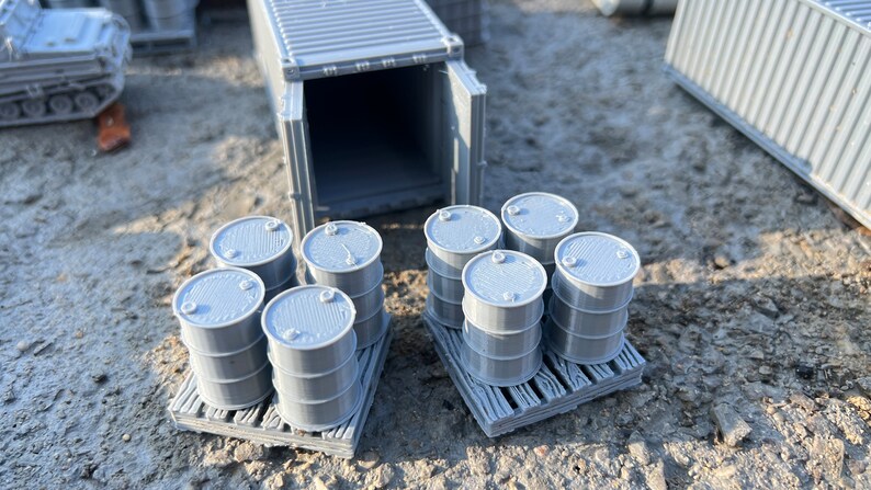 May include: Two pallets of gray plastic barrels, each pallet holding six barrels. The barrels are stacked two high. A gray shipping container with its doors open is in the background.