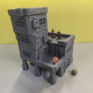 May include: A gray, multi-level miniature building with a futuristic design, featuring rectangular windows and air conditioning units. Two miniature figures stand on the rooftop. A red and a green die are also present, suggesting a tabletop game setting.