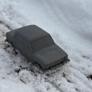 May include: A gray toy car sits in the snow. The miniature vehicle is a sedan style, with visible details like windows and wheels. The car is surrounded by fresh, white snow, creating a winter scene.