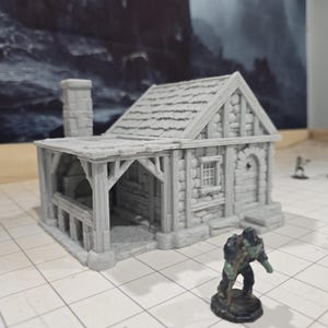 May include: A gray 3D printed miniature stone cottage with a wooden porch and a chimney. The cottage has a small window and a door. The miniature is sitting on a gray tiled surface.