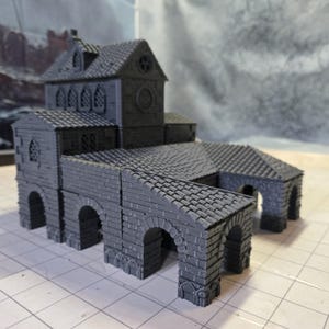 May include: A gray 3D printed model of a medieval building with a tiled roof, arched doorways, and a clock tower. The building has a stone facade and is designed for tabletop gaming.