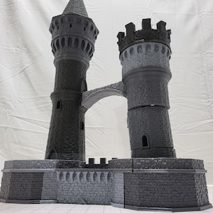 May include: A gray 3D printed castle model with two towers connected by a bridge. The towers have detailed brickwork and windows. The castle is set on a base with a stone wall.