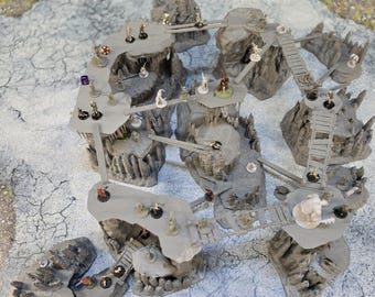 May include: A detailed tabletop gaming terrain set, featuring multiple gray rock formations connected by bridges and walkways. Miniature figures are placed on the platforms, suggesting a role-playing game setting. The set is designed for fantasy or adventure games.
