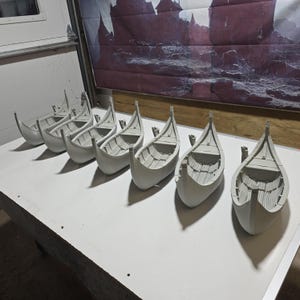 May include: A set of seven unpainted light gray miniature boat models arranged in a row. Each vessel features a curved hull, a pointed bow and stern, and detailed interior planking textures, representing a classic longship design for tabletop gaming.