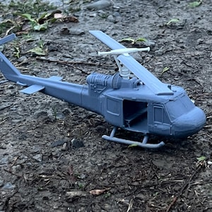 May include: A gray plastic model of a helicopter with a white rotor blade. The helicopter is sitting on a bed of gravel.